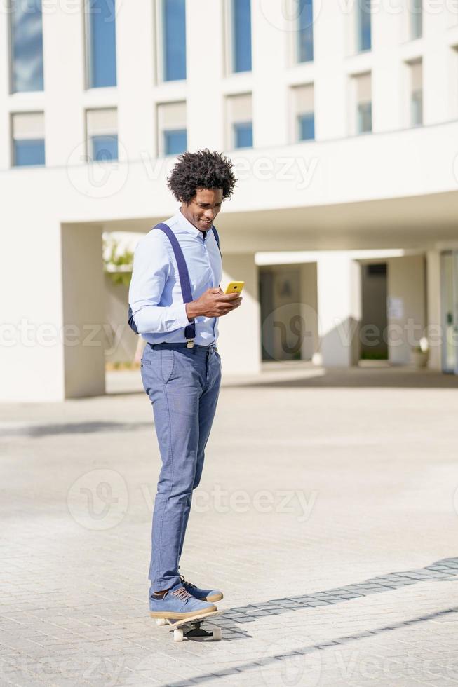 uomo d'affari nero su uno skateboard guardando il suo smartphone all'aperto. foto