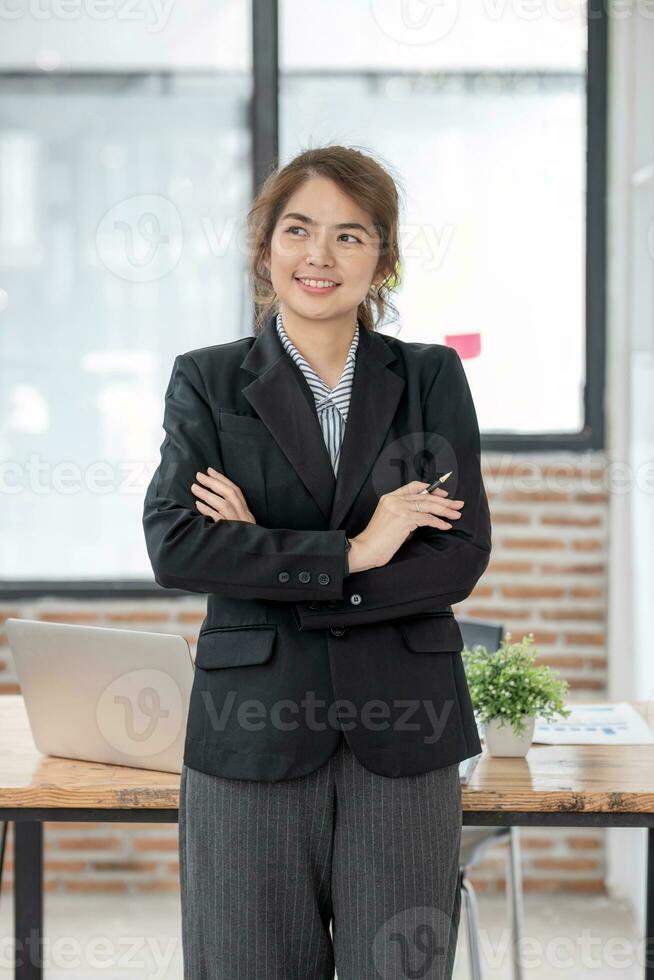 ritratto, giovane fiducioso sorridente asiatico attività commerciale donna indossare completo da uomo in piedi nel ufficio con braccia incrociato. foto