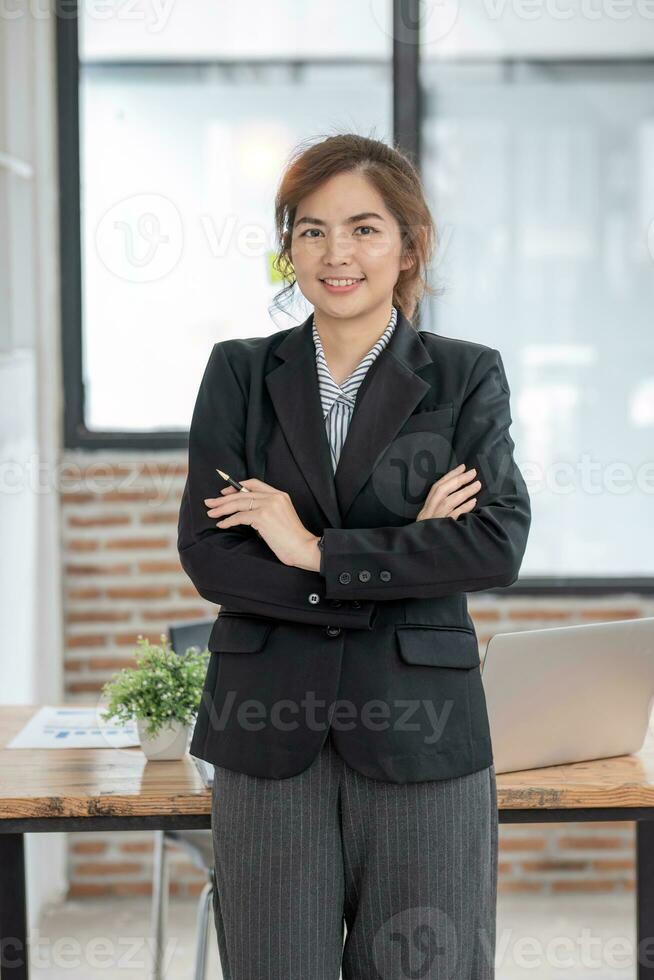 ritratto, giovane fiducioso sorridente asiatico attività commerciale donna indossare completo da uomo in piedi nel ufficio con braccia incrociato. foto