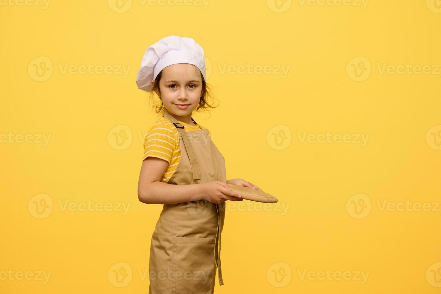 autentico poco ragazza nel dello chef cappello e grembiule, sorrisi a telecamera, Tenere un' di legno asse, isolato giallo studio fondale foto