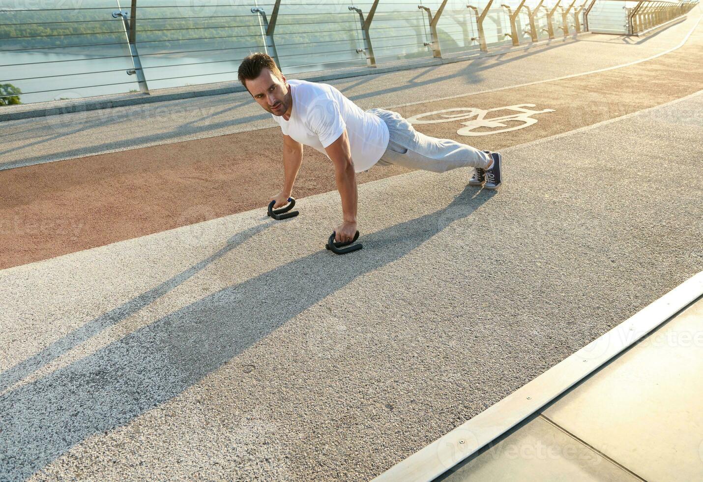 pieno lunghezza ritratto di un' bello mezzo anziano europeo uomo, muscolare costruire atleta fare spingere UPS durante un all'aperto allenarsi su il città ponte presto nel il mattina foto