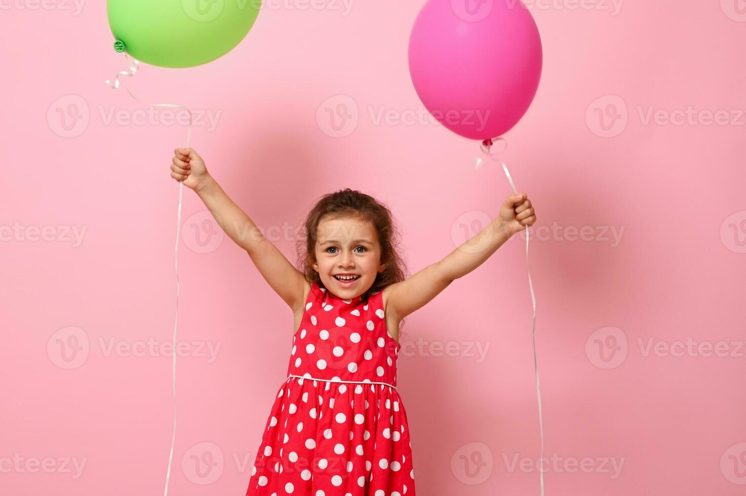 bambino ragazza vestito nel rosa vestito con polka puntini modello raccolta sua braccia su con colorato palloncini nel sua mani, sorridente guardare a il telecamera, isolato su rosa sfondo con copia spazio foto