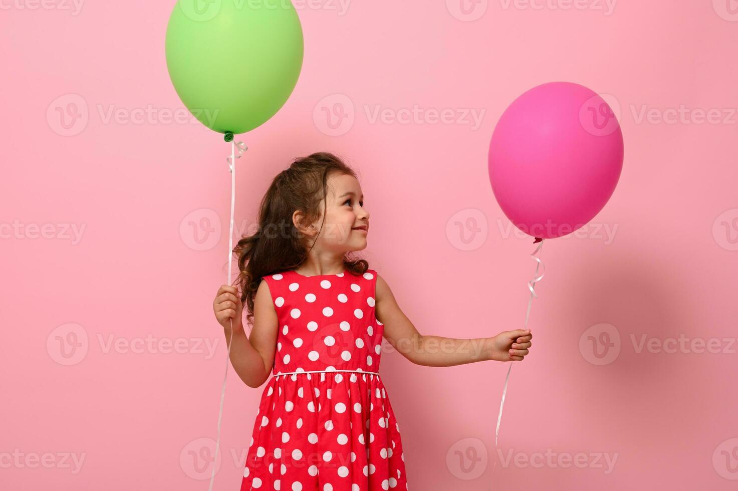 bellissimo bellissima 4 anno bella compleanno ragazza nel rosa polka punto vestire, detiene Due multicolore palloncini, gioisce guardare a un' verde Palloncino nel sua mano, isolato su rosa sfondo con copia spazio. foto