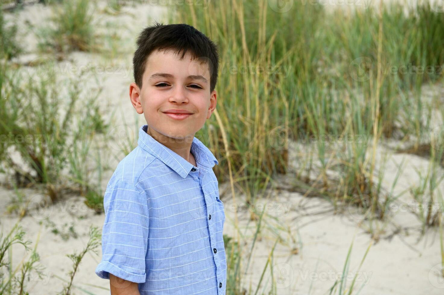 avvicinamento di il viso di un' carino sorridente adolescenziale ragazzo in piedi contro il fondale di steppa boschetti e sabbioso suolo. bello bambino carino sorrisi guardare a il telecamera. colpo alla testa di adorabile ragazzo foto