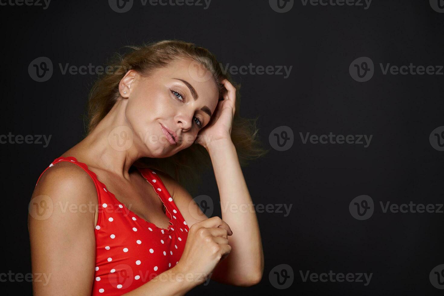 attraente europeo donna con salutare pulito fresco raggiante abbronzato pelle e biondo brillante dritto capelli, naturale trucco, indossare rosso costume da bagno con bianca polka puntini isolato su nero sfondo foto