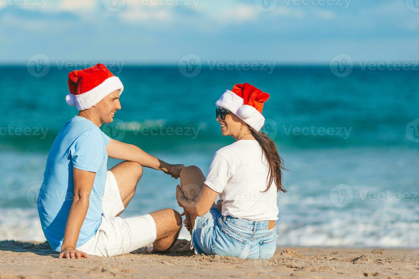giovane romantico coppia nel rosso Santa cappelli seduta su tropicale bianca sabbia spiaggia festeggiare Natale foto