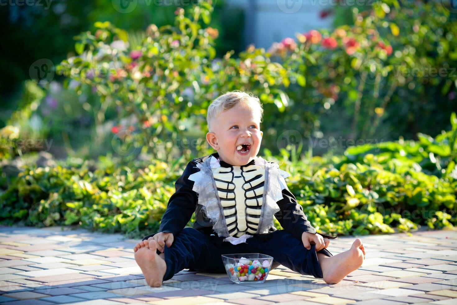 un' poco ragazzo si siede su il erba nel un' scheletro completo da uomo e mangia dolci. Halloween vacanza. foto