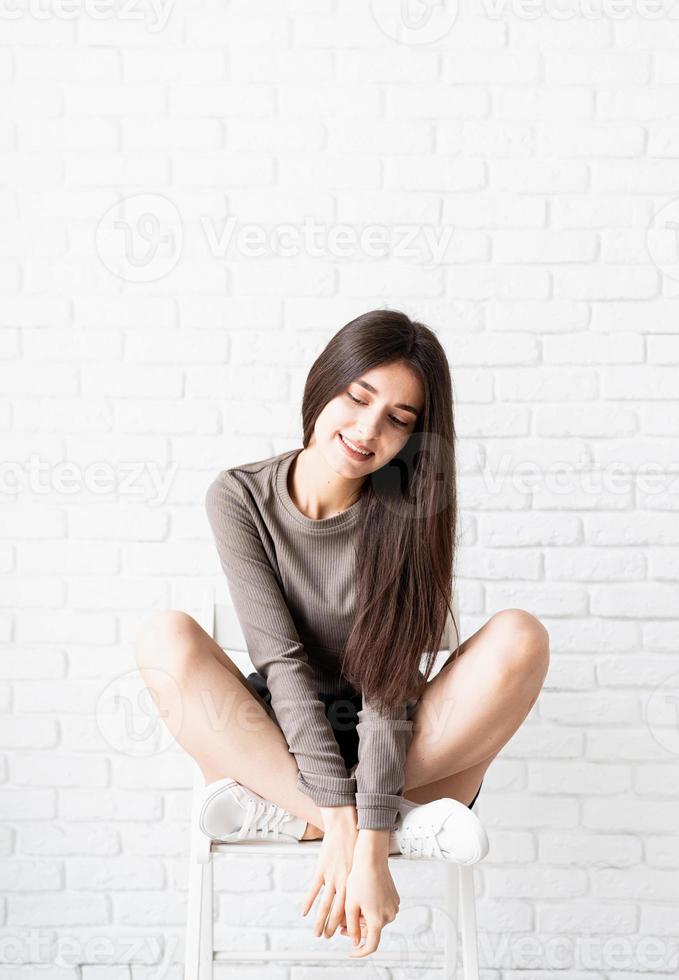 donna con i capelli lunghi che si siede sul fondo del muro di mattoni bianchi, pensando foto