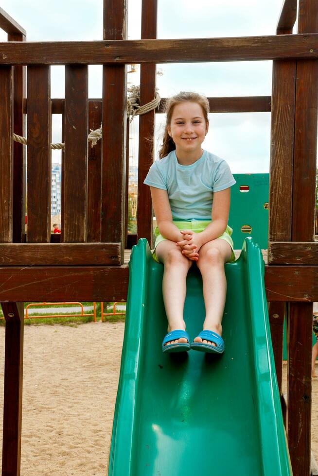 un' poco ragazza su il terreno di gioco si siede su un' diapositiva e sorrisi allegramente foto