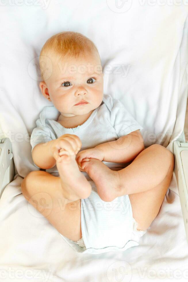 carino poco neonato ragazza con divertente viso guardare a telecamera su bianca sfondo. infantile bambino riposo giocando dire bugie giù su culla letto a casa. maternità contento bambino concetto. foto