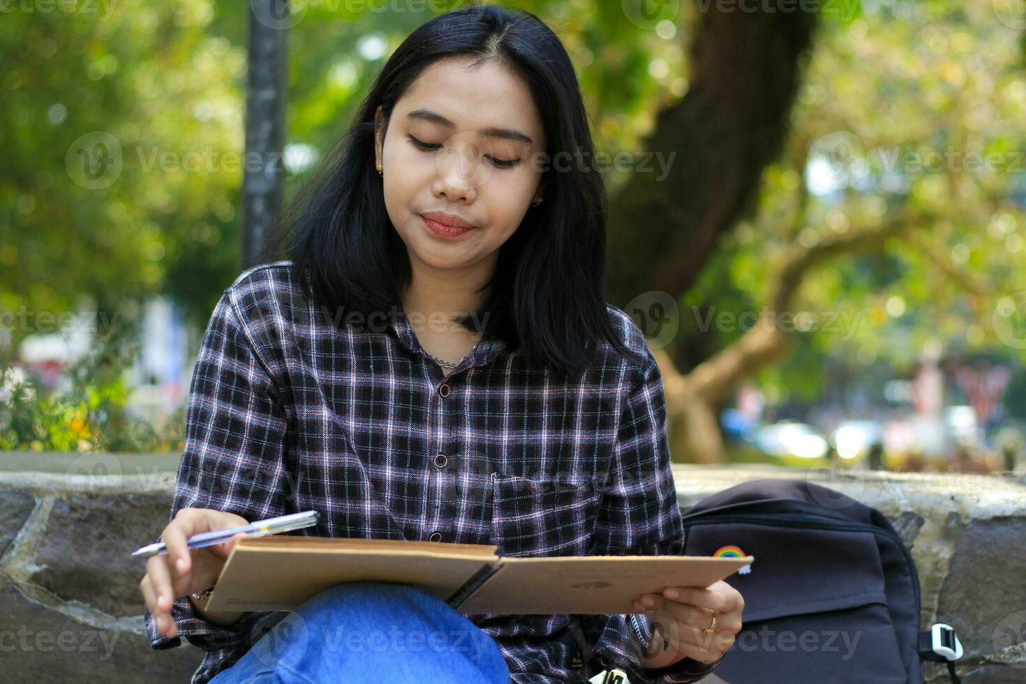 bellissimo asiatico giovane donna Università alunno focalizzata scrittura su taccuino e lettura libro nel all'aperto città parco foto