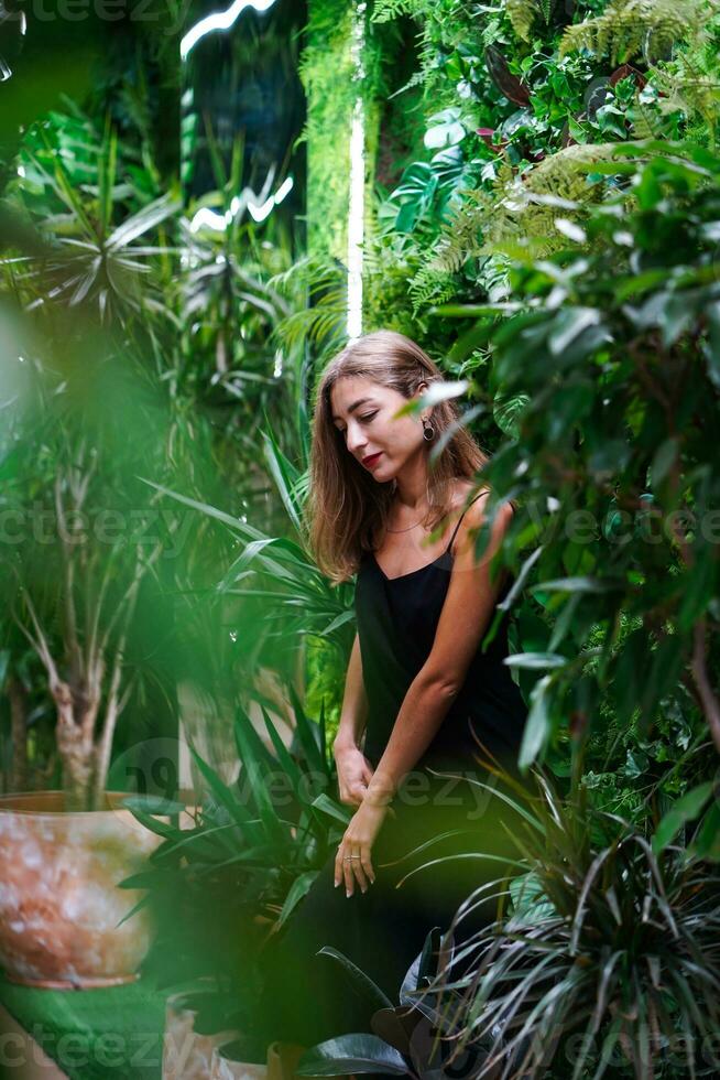 ragazza guardare giù pensieroso. foresta pluviale. giovane donna indossare nero vestito su magro cinghie sta tra verde impianti dentro disposizione. giovane bellissimo donna modello pose tra verde tropicale impianti. foto