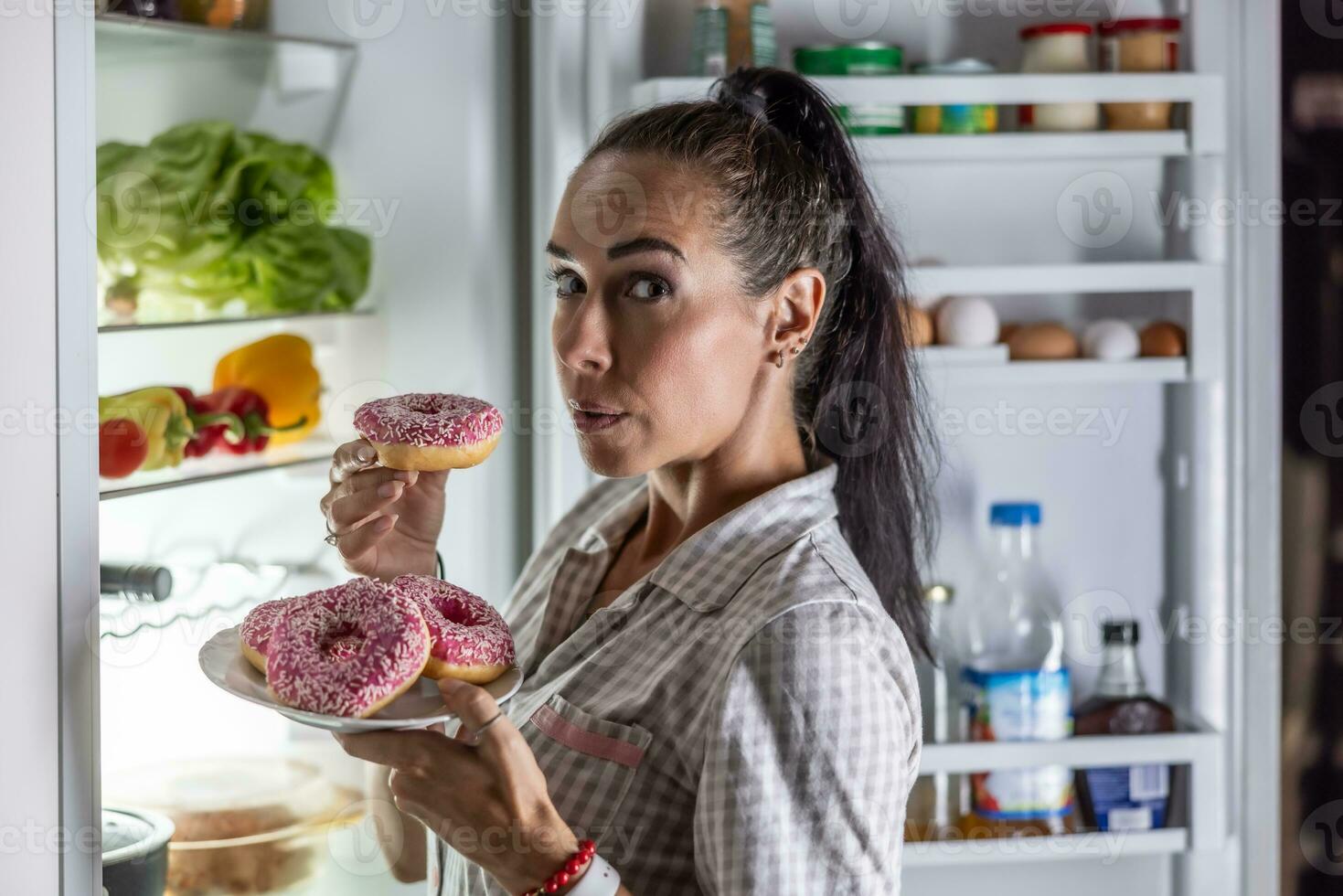 un' giovane Affamato donna è di per mangiare dolce ciambelle di il Aperto frigorifero in ritardo nel il sera foto