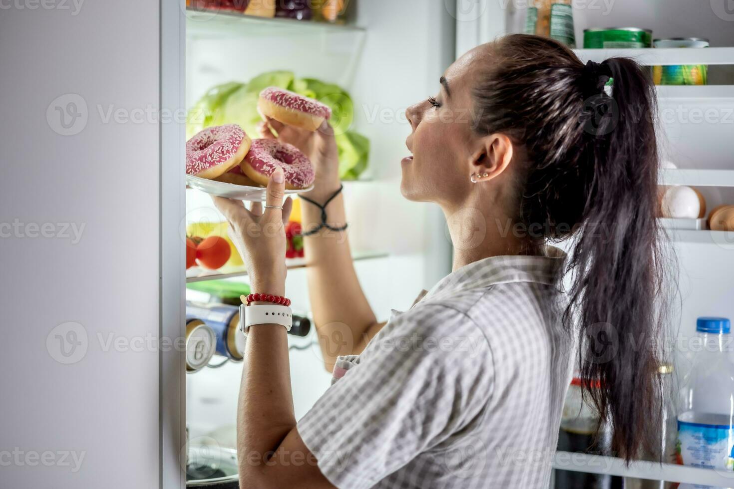 un' giovane Affamato donna è di per mangiare dolce ciambelle di il Aperto frigorifero in ritardo nel il sera foto