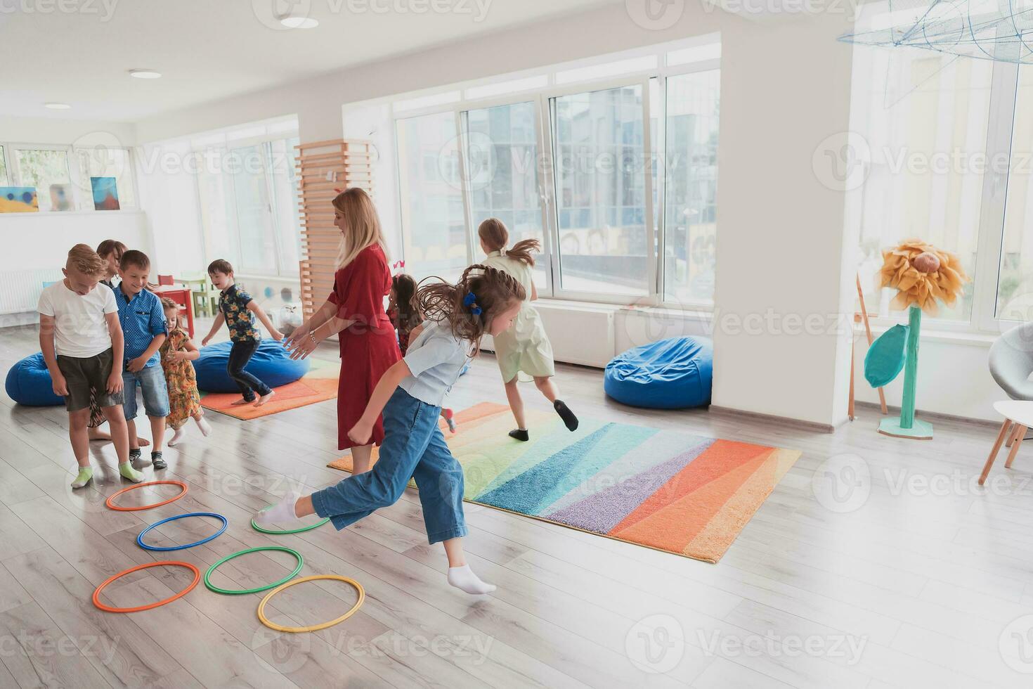 piccolo asilo scuola bambini con femmina insegnante su pavimento in casa nel aula, fare esercizio. salto al di sopra di hula cerchio cerchi traccia su il pavimento. foto