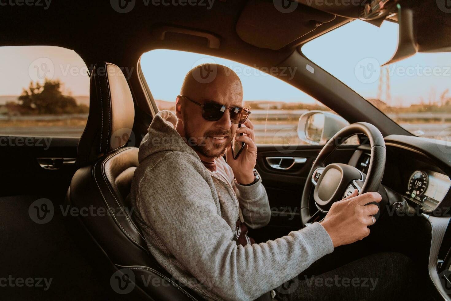 un' uomo con un' occhiali da sole guida un' auto e parlando su smartphone a tramonto. il concetto di auto viaggio foto