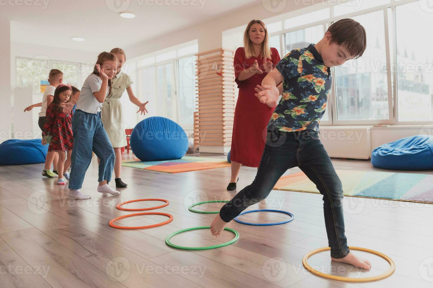 piccolo asilo scuola bambini con femmina insegnante su pavimento in casa nel aula, fare esercizio. salto al di sopra di hula cerchio cerchi traccia su il pavimento. foto