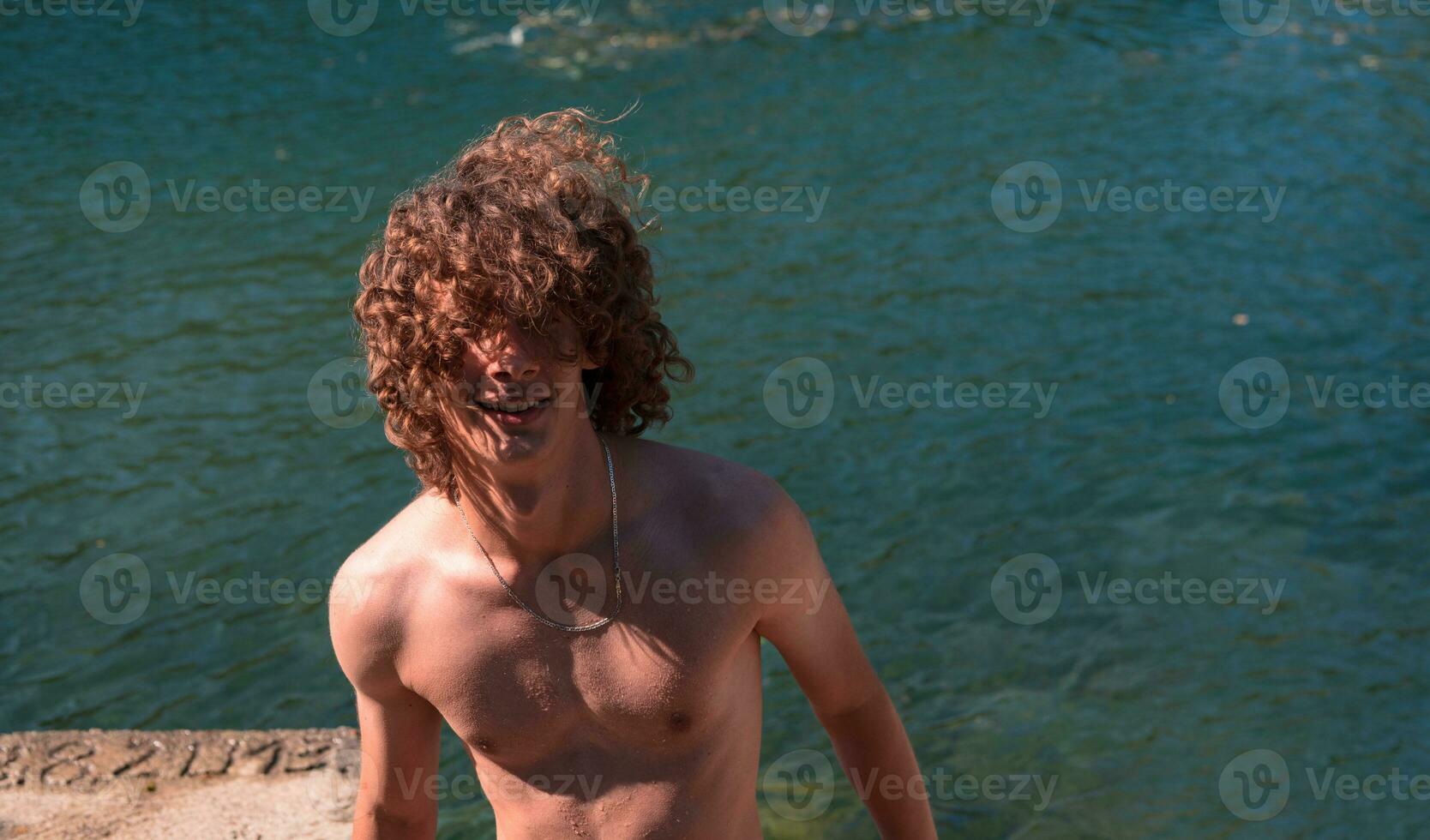 ritratto di un' giovane adolescente ragazzo con Riccio bagnato capelli vicino il fiume avendo divertimento con amici a un' estate festa. foto