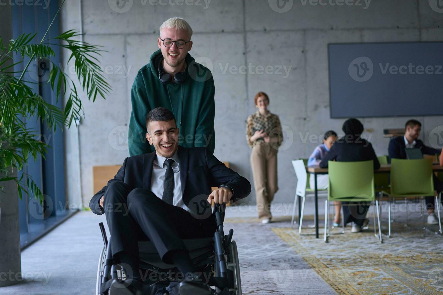 un' giovane imprenditore e il suo collega, chi è un' persona con un' disabilità, divertimento insieme attraverso un' di grandi dimensioni, moderno ufficio, dimostrando il energia di lavoro di squadra, inclusività, e determinazione come essi guidare il sedia a rotelle foto