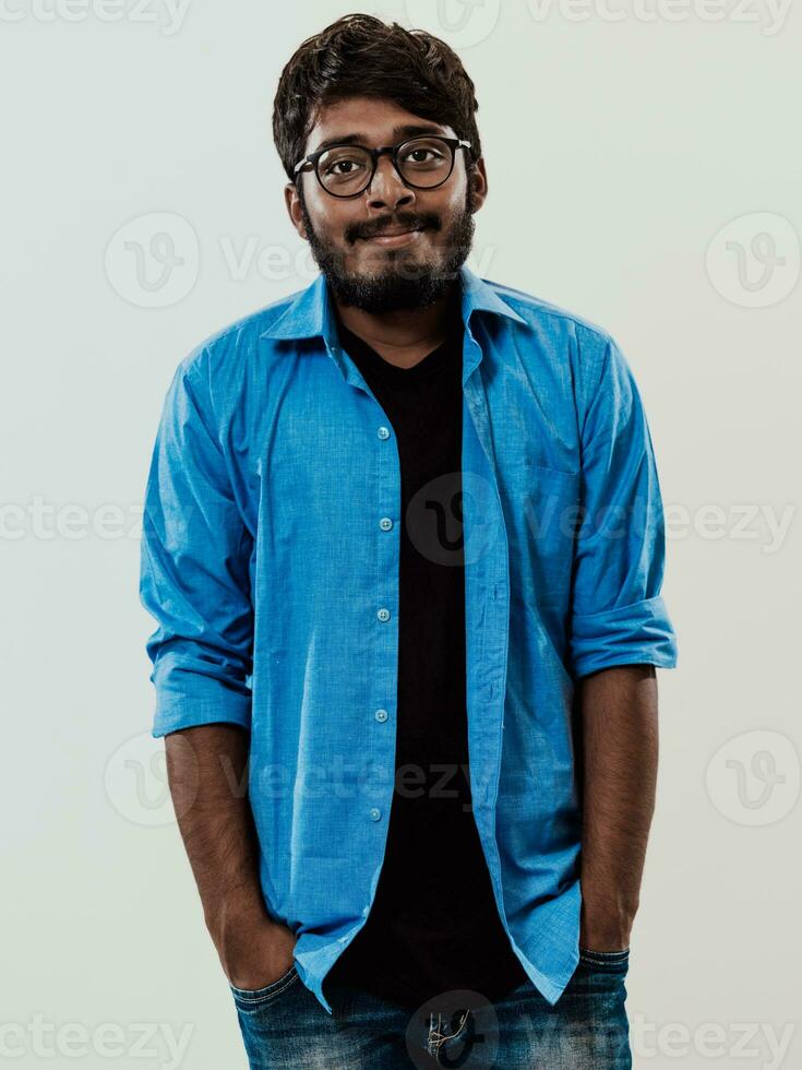 indiano sorridente giovane uomo con blu camicia e bicchieri in posa su grigio sfondo foto