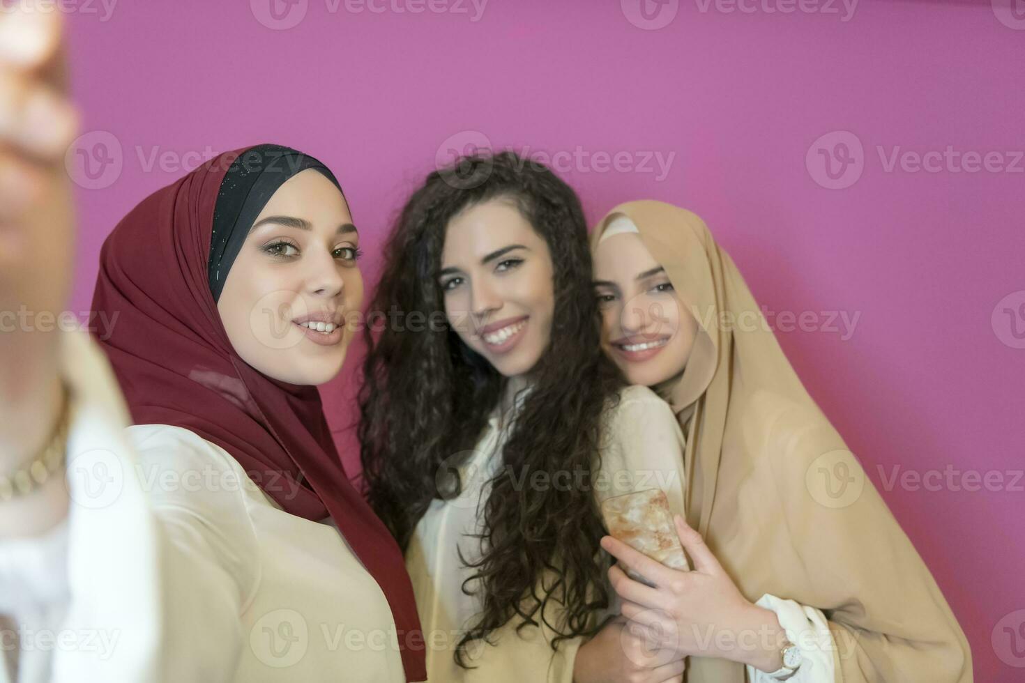 gruppo di bellissimo musulmano donne Due di loro nel alla moda vestito con hijab utilizzando mobile Telefono mentre assunzione autoscatto immagine isolato su rosa sfondo che rappresentano moderno Islam moda tecnologia foto