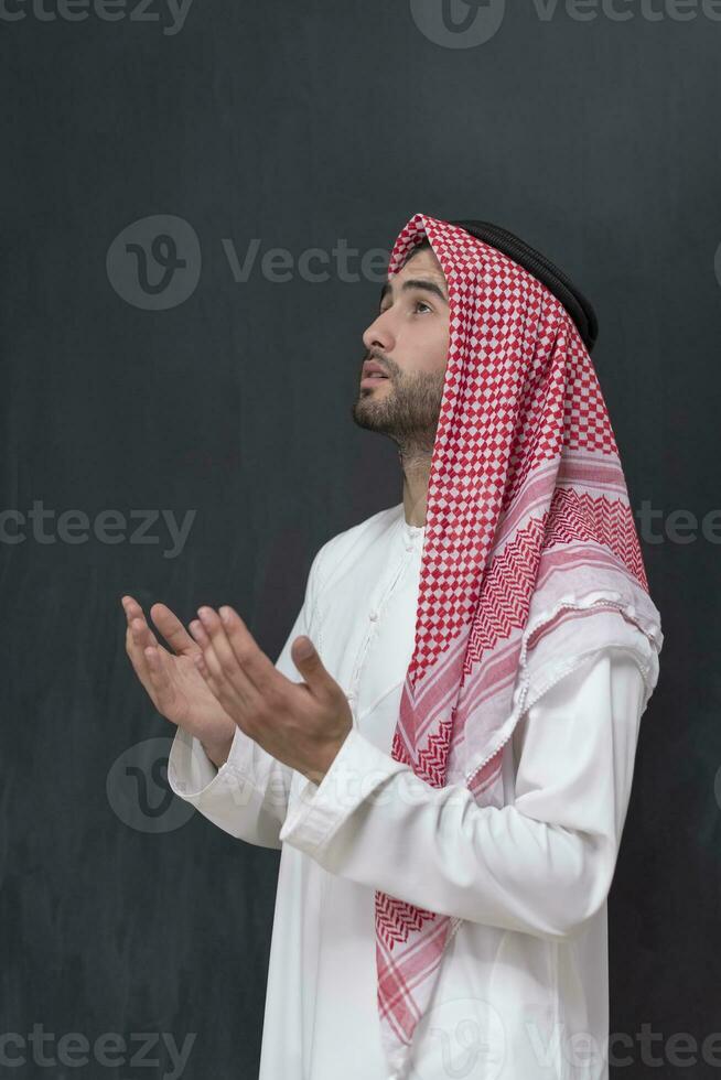 un' giovane arabo uomo nel tradizionale Abiti fabbricazione un' tradizionale preghiera per Dio mantiene il suo mani nel preghiere gesto nel davanti di un' nero sfondo foto