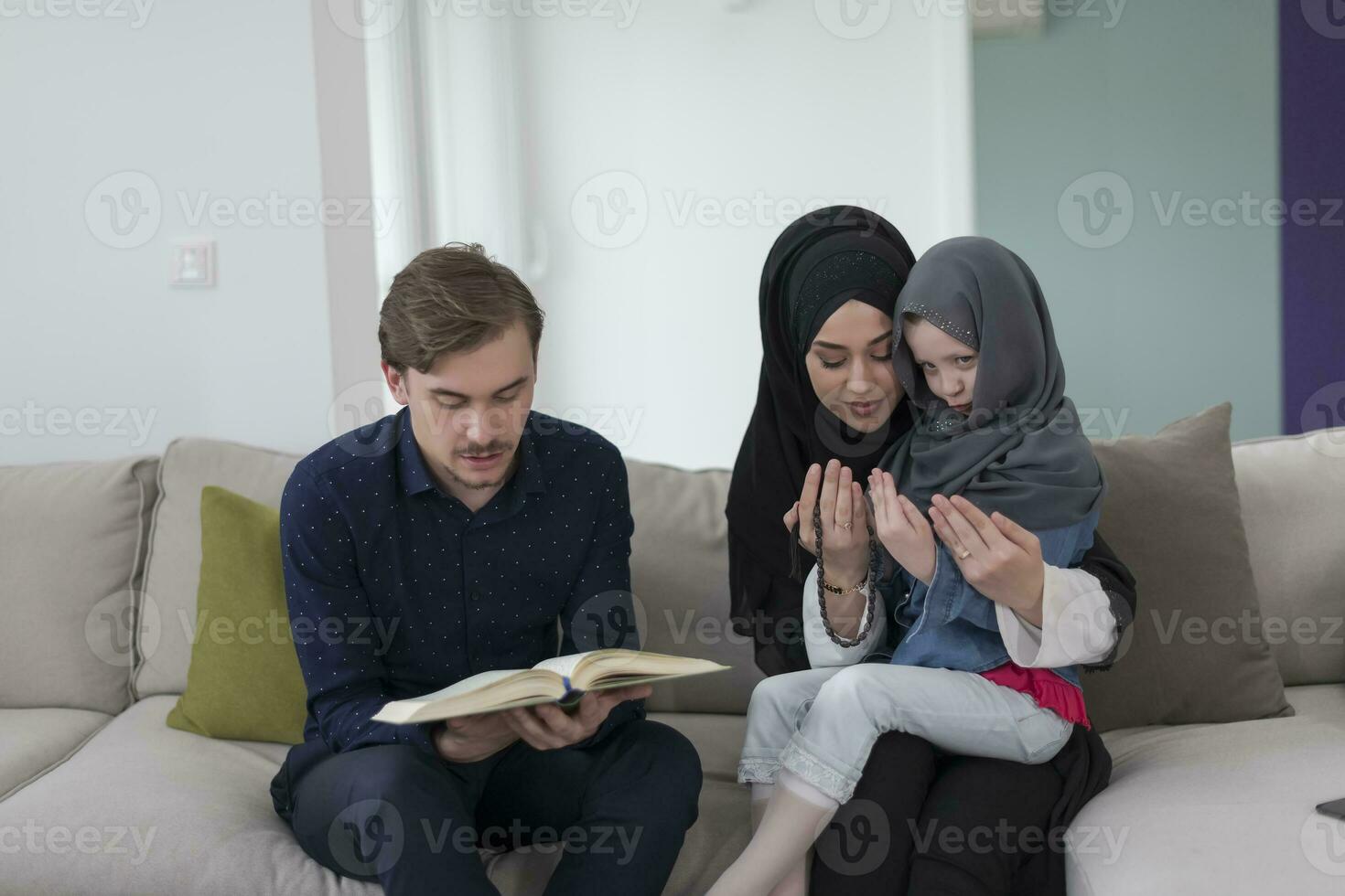 tradizionale musulmano famiglia genitori con bambini lettura Corano e preghiere insieme su il divano prima iftar cena durante un' Ramadan festa a casa foto