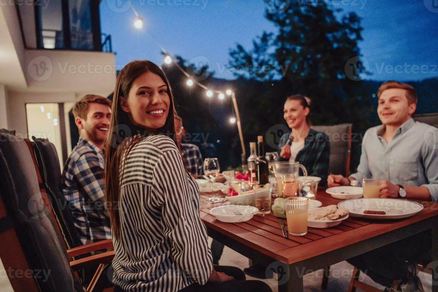un' gruppo di giovane diverso persone avendo cena su il terrazza di un' moderno Casa nel il sera. divertimento per amici e famiglia. celebrazione di vacanze, matrimoni con barbecue. foto