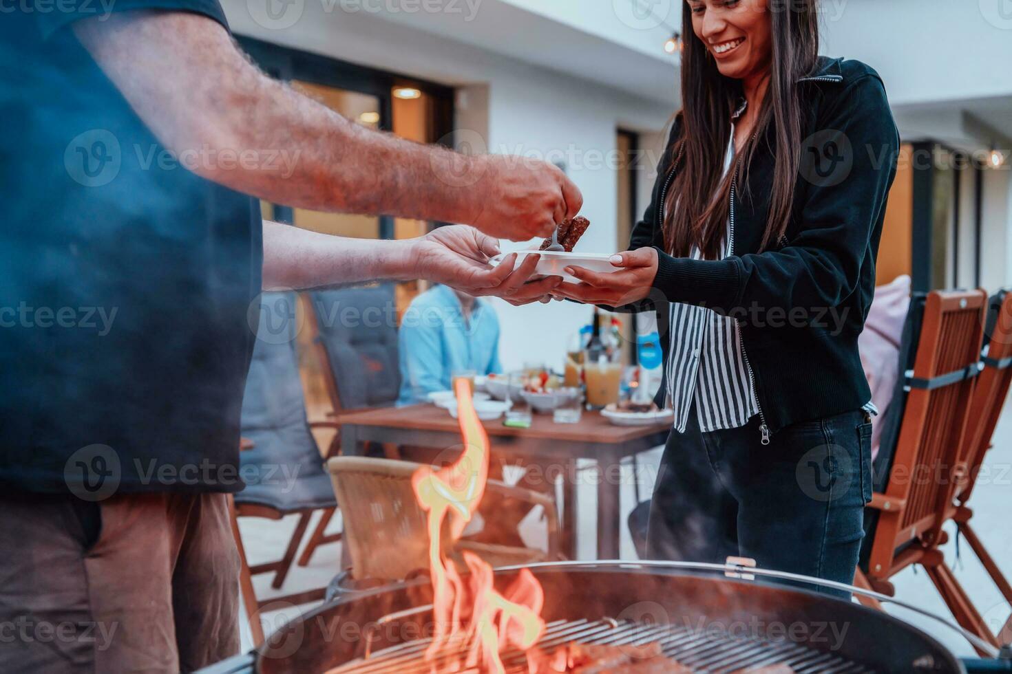 un' gruppo di amici e famiglia barbecue insieme nel il sera su il terrazza nel davanti di un' grande moderno Casa foto