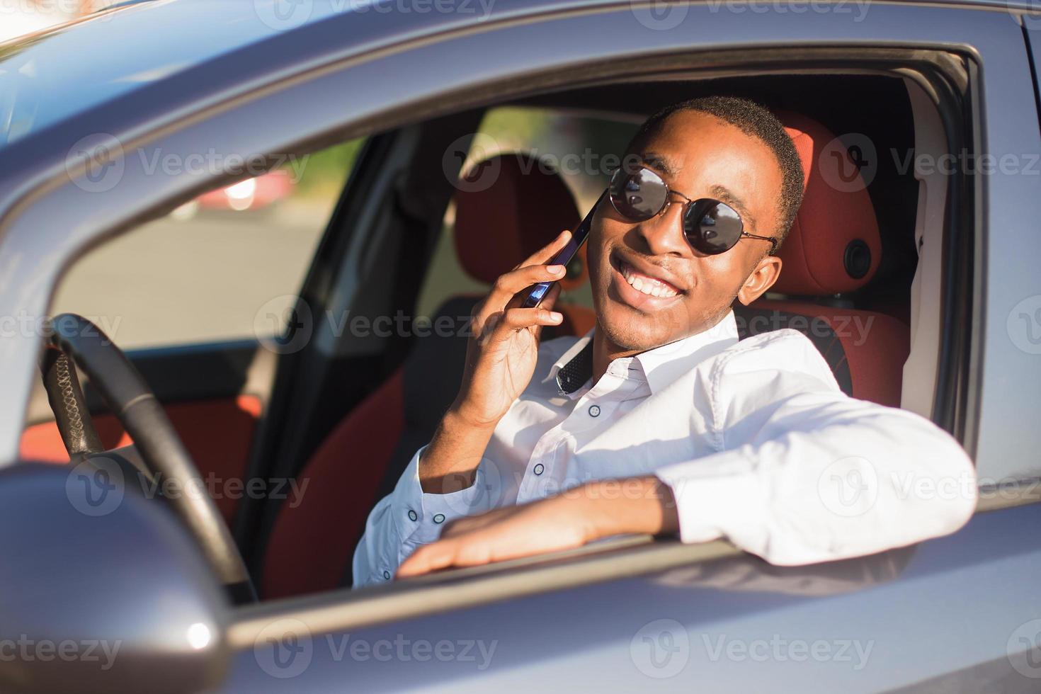 felice afroamericano alla guida di un'auto con un telefono, in estate foto