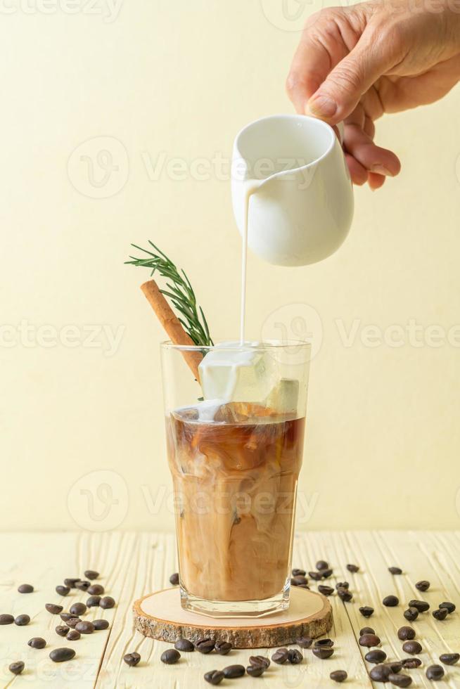 versando il latte in un bicchiere di caffè nero con cubetto di ghiaccio, cannella e rosmarino su fondo di legno foto