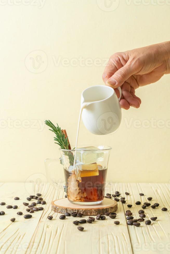 versando il latte in un bicchiere di caffè nero con cubetto di ghiaccio, cannella e rosmarino su fondo di legno foto