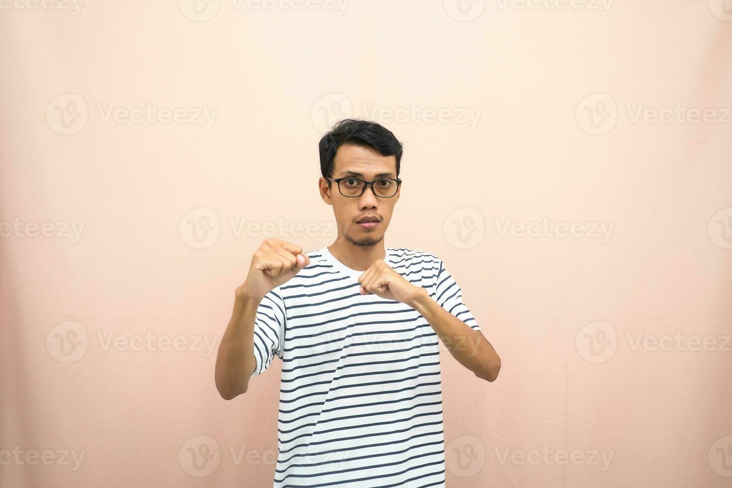 asiatico uomo con bicchieri indossare casuale a strisce camicia, cazzotto cazzotto posa, indicando forte o pronto per combattere. isolato beige sfondo. foto