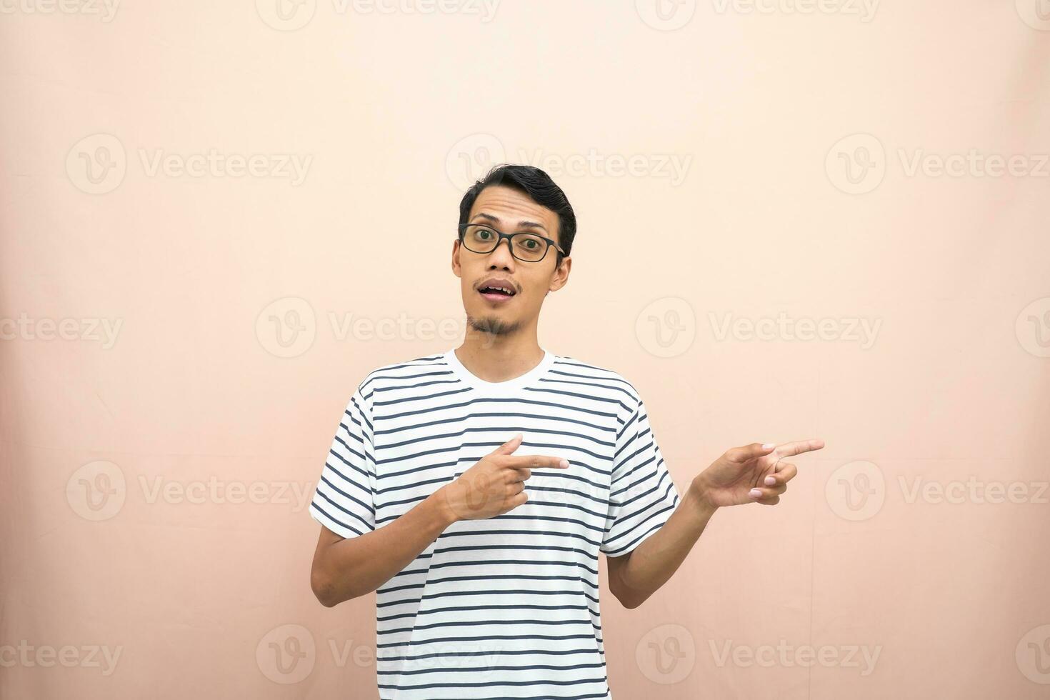 asiatico uomo con bicchieri indossare casuale a strisce camicia, sussurrando posa mentre puntamento per il lato. isolato beige sfondo. foto