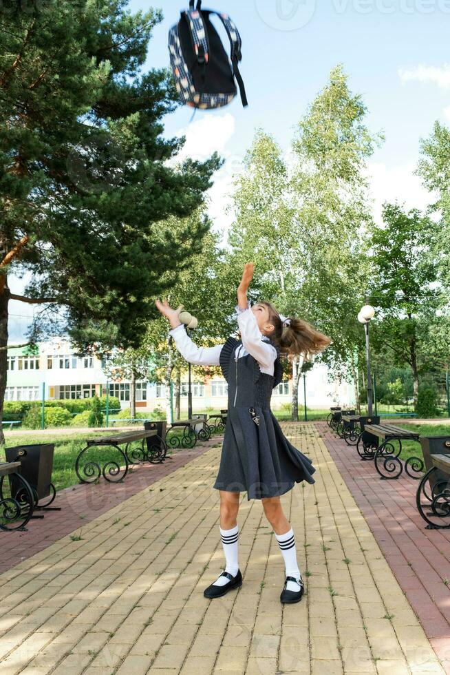 allegro divertente ragazza con sdentato Sorridi nel scuola uniforme con bianca fiocchi nel scuola cortile lanci il zaino. indietro per scuola, settembre 1. contento allievo. primario formazione scolastica, elementare classe. foto