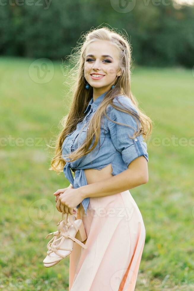 sorridente giovane donna nel casuale Abiti passeggiate attraverso il parco. lei è Tenere scarpe nel sua mani. lei è gratuito e contento. il concetto di la libertà e gioventù foto