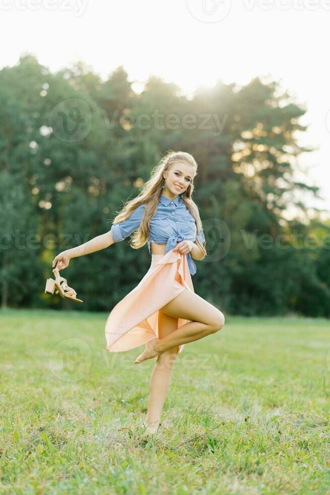 sorridente giovane donna nel casuale Abiti passeggiate attraverso il parco. lei è Tenere scarpe nel sua mani. lei è gratuito e contento. il concetto di la libertà e gioventù foto