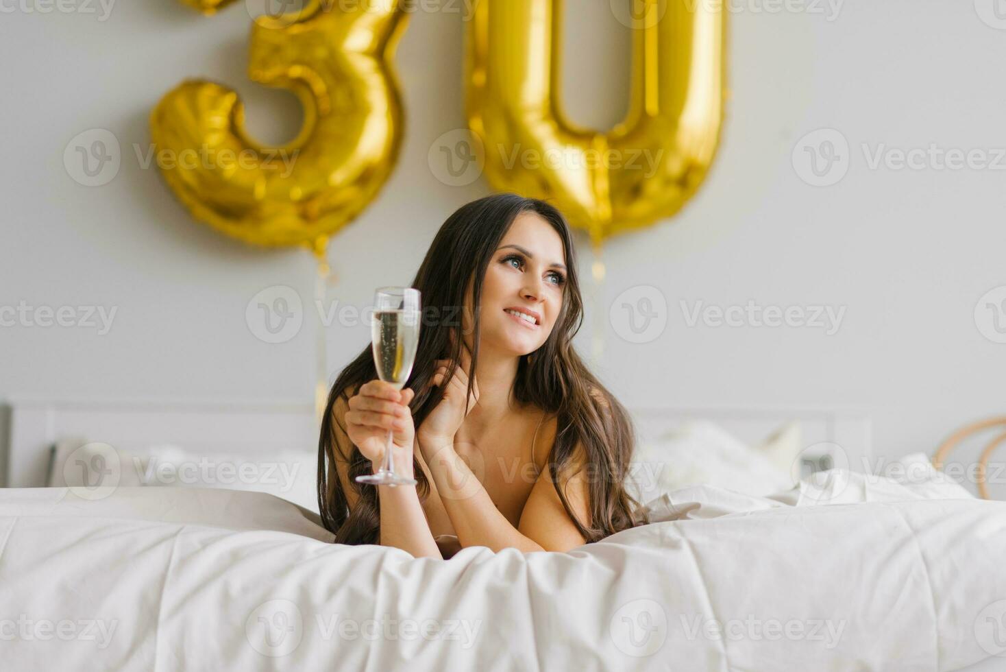 bellissimo donna svegliato su nel letto nel il mattina e celebra sua anni trenta con un' bicchiere di Champagne nel un' camicia da notte foto