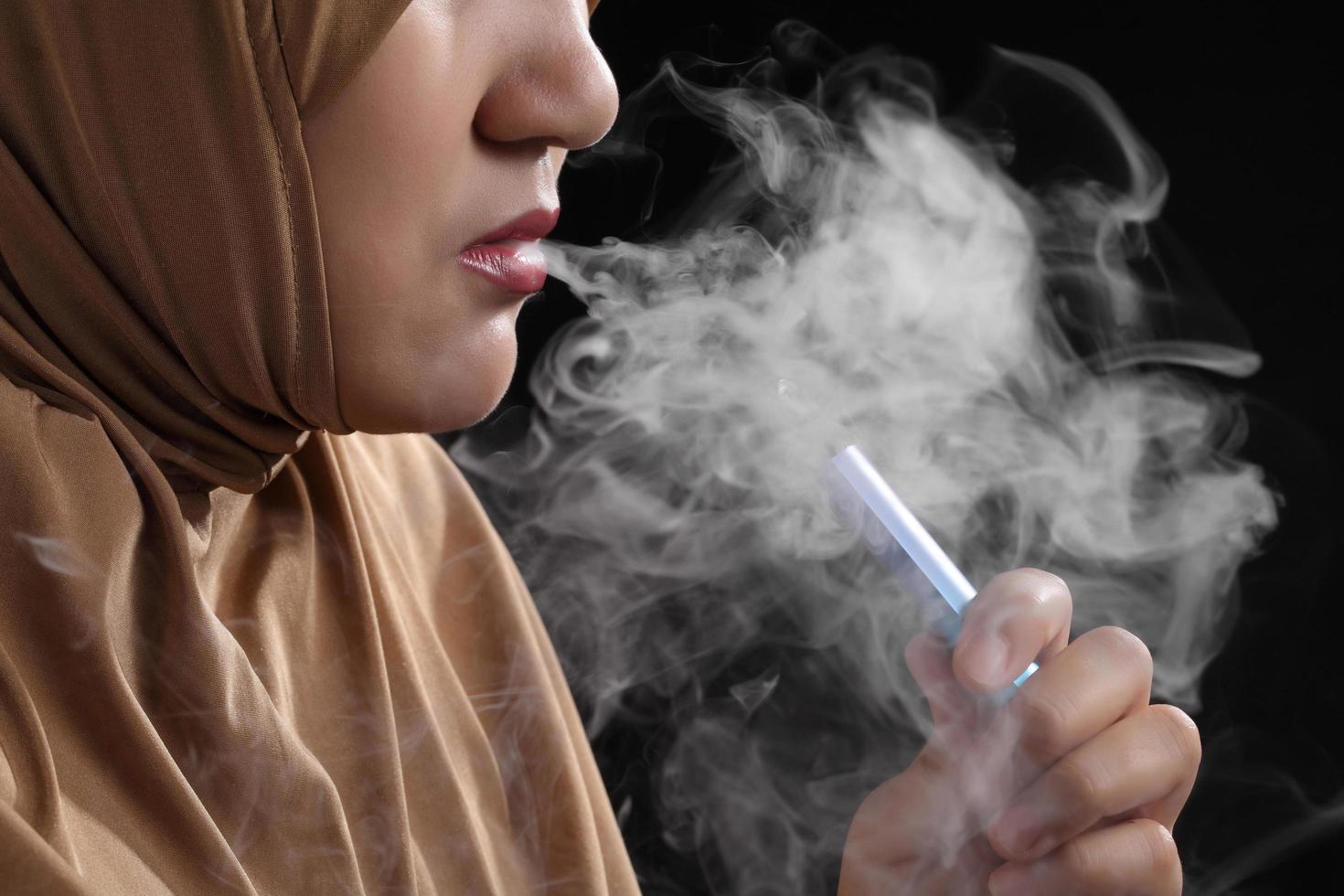 Close up giovane donna musulmana che fuma sigaretta elettronica su sfondo nero foto