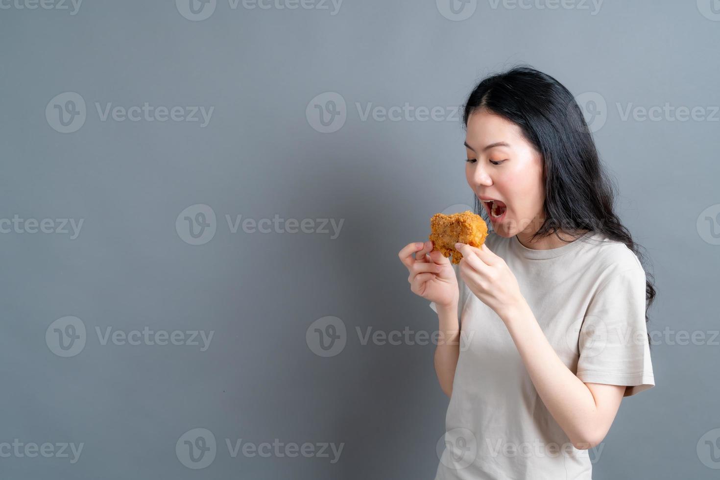 giovane donna asiatica che indossa t-shirt con faccia felice e si diverte a mangiare pollo fritto su sfondo grigio foto