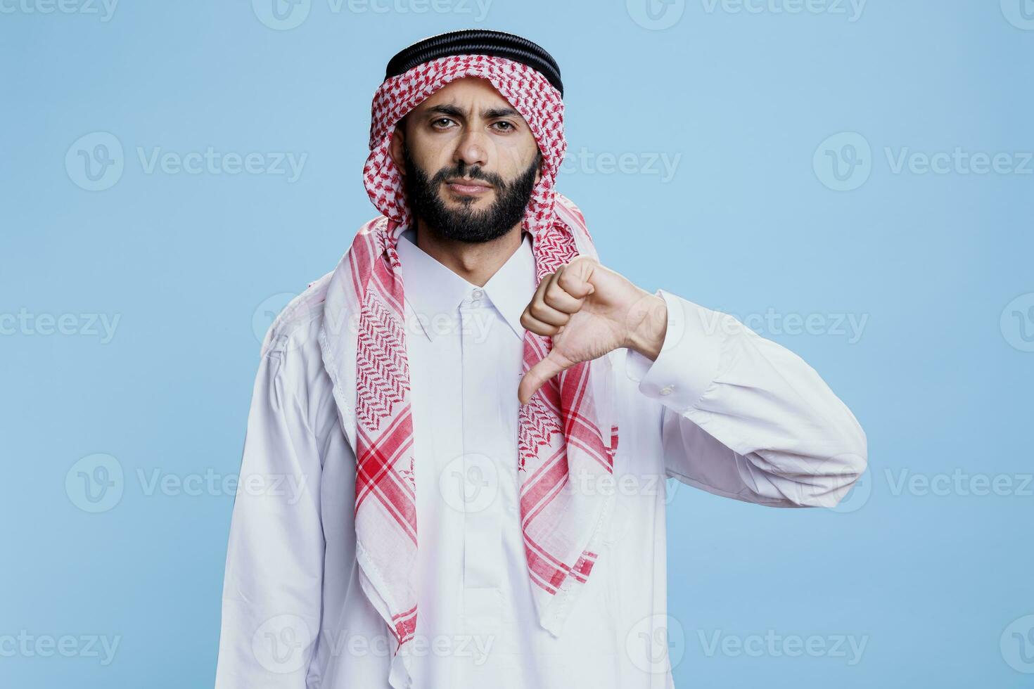 grave arabo uomo indossare tradizionale Abiti in posa con pollice giù gesto studio ritratto. musulmano persona nel bianca islamico accappatoio e foulard mostrando disaccordo gesto e guardare a telecamera foto