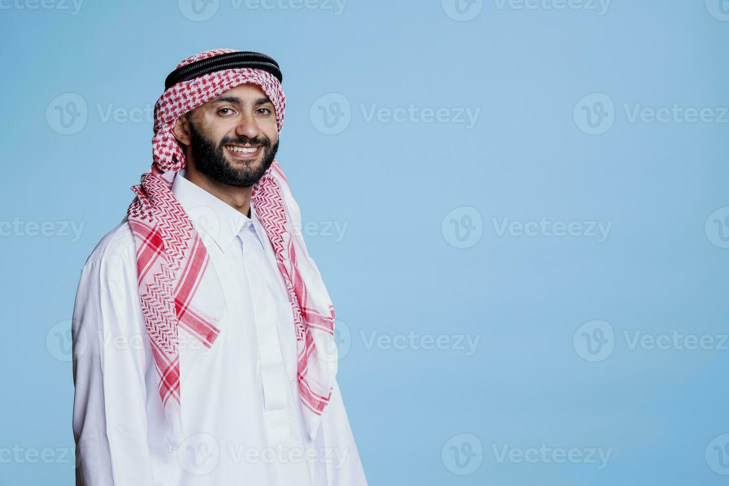 allegro musulmano uomo con spensierato espressione vestito nel tradizionale arabo Abiti guardare a telecamera. sorridente persona indossare scacchi rosso e bianca copricapo studio ritratto foto