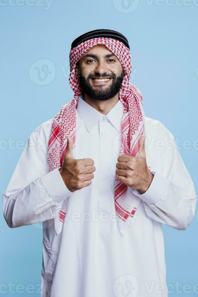 sorridente musulmano uomo vestito nel tradizionale islamico Abiti mostrando Due pollici su ritratto. arabo fabbricazione approvazione gesto con dita e guardare a telecamera con positivo espressione foto