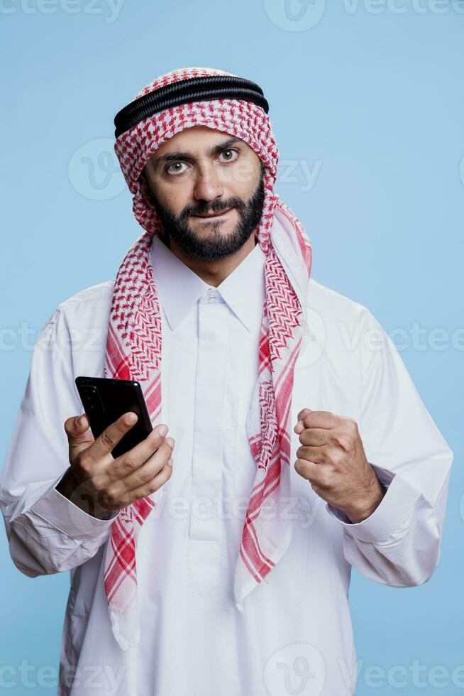 eccitato musulmano uomo indossare tradizionale Abiti utilizzando smartphone, mostrando sì gesto con stretto cazzotto ritratto. arabo vestito nel thobe e ghutra Tenere mobile Telefono e in posa con vincitore concetto foto