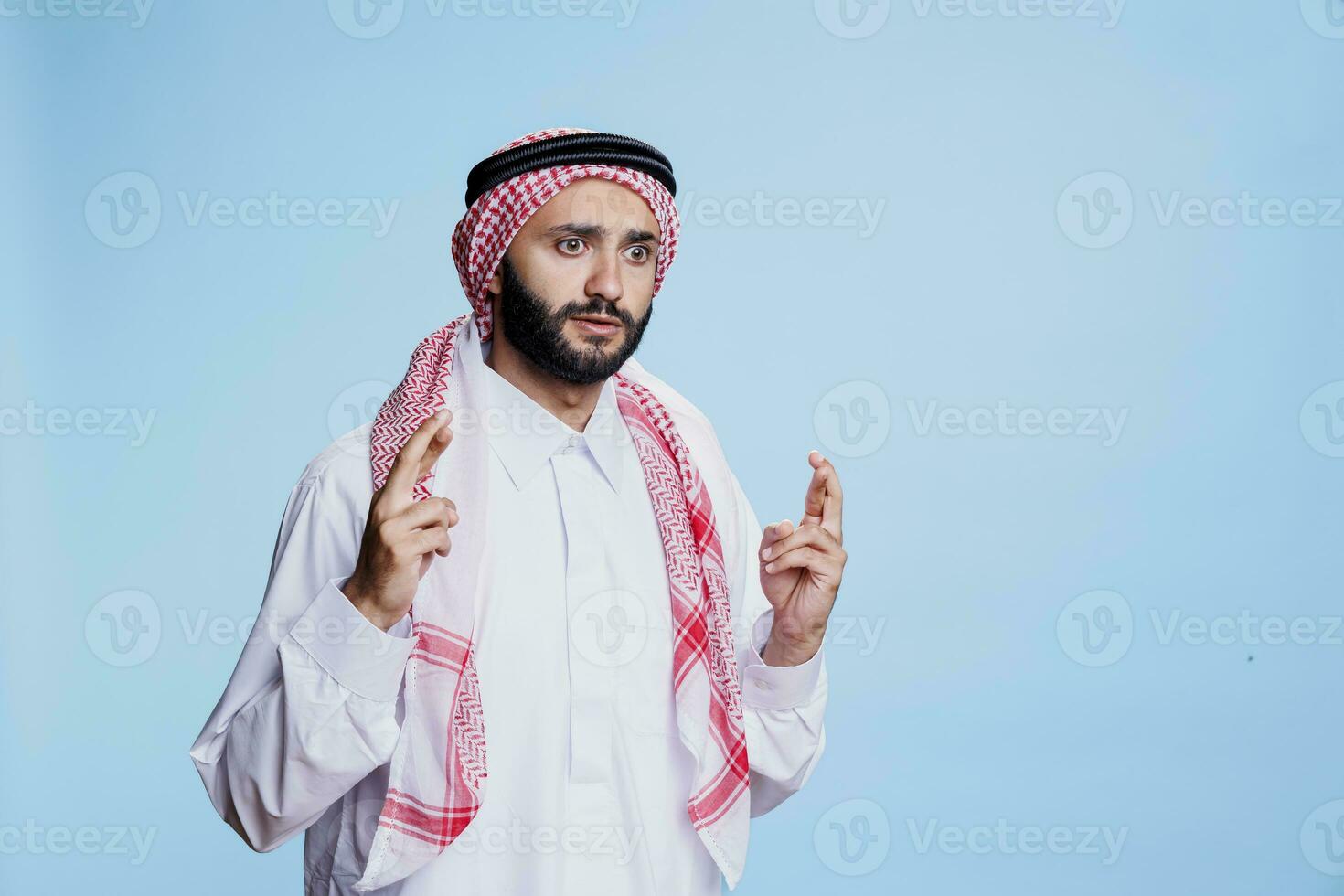 impaurito uomo vestito nel culturale musulmano Abiti in piedi con attraversato dita. terrorizzato arabo persona con occhi largo Aperto in posa con superstizioso gesto per fortuna nel studio su blu sfondo foto