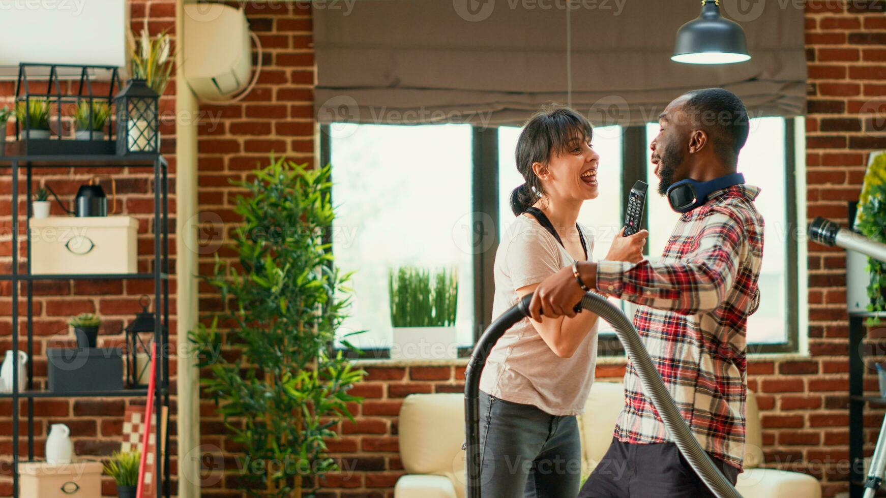 diverso coppia avendo divertimento pulizia vivente camera con vuoto addetto alle pulizie e Mocio, cantando e ascoltando per musica. giovane contento partner godendo primavera pulizia, passare l'aspirapolvere appartamento piani. foto