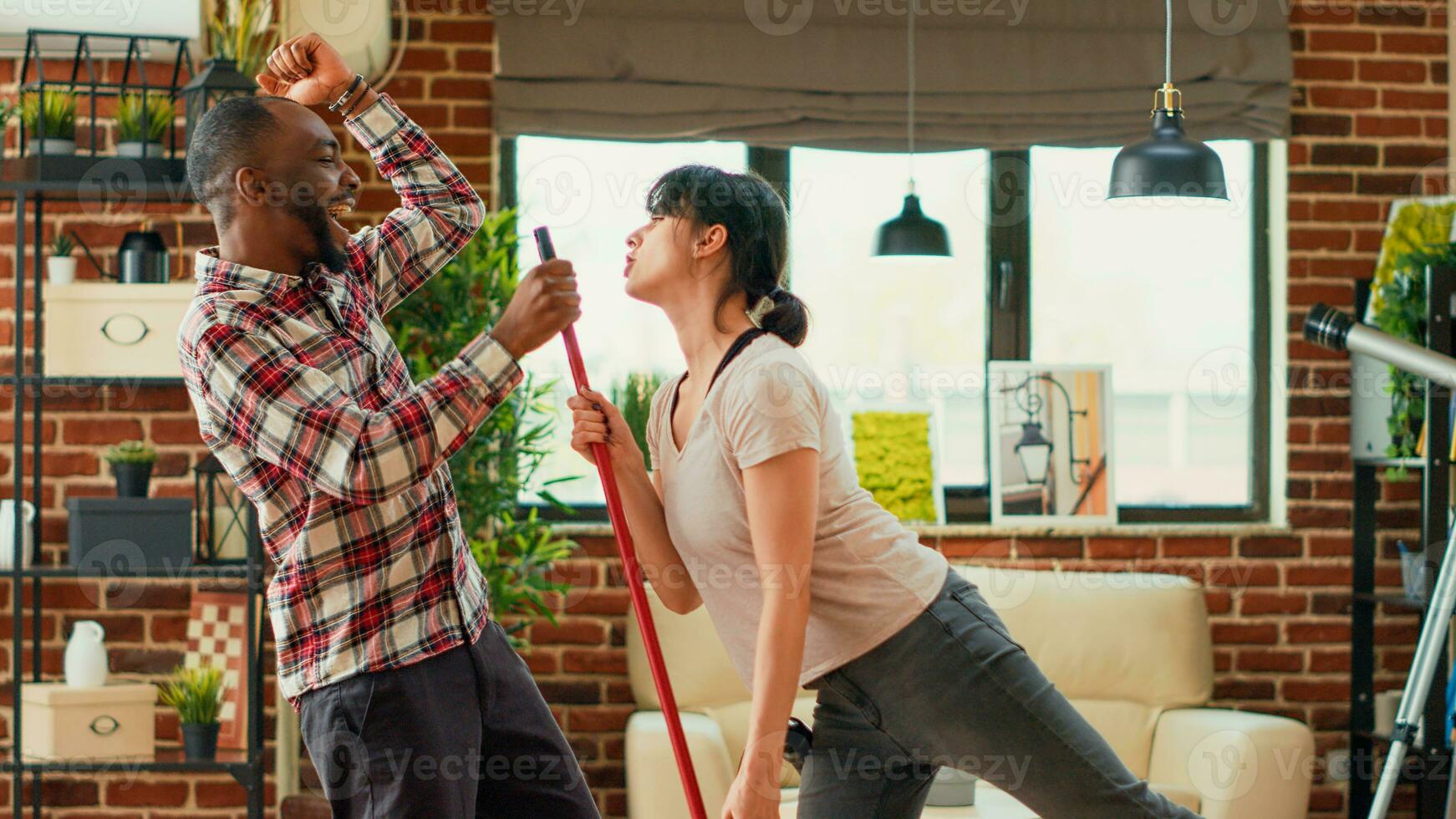 moderno sorridente coppia danza e pulizia appartamento, rastrellamento e passare l'aspirapolvere vivente camera di legno piani. giovane positivo partner avendo divertimento con danza si sposta, godendo lavori di casa. palmare sparo. foto