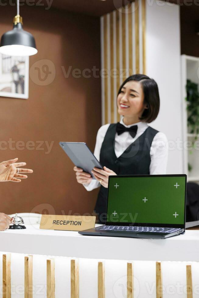 addetto alla reception avendo schermo verde su scrivania, accogliente ospiti a Hotel e di partenza dai un'occhiata nel processi con tavoletta. asiatico donna utilizzando il computer portatile mostrando isolato Schermo con croma chiave copyspace. foto