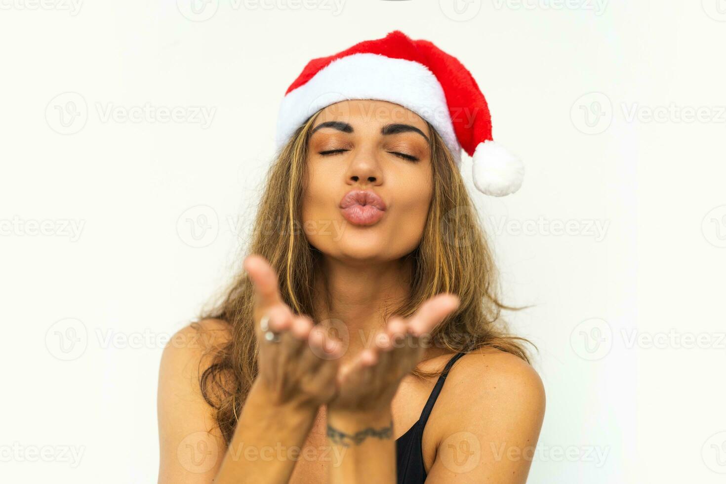 bella donna con nel Santa cappello e elegante nero vestito Spedire bacio per telecamera e in posa su bianca sfondo nel studio. foto