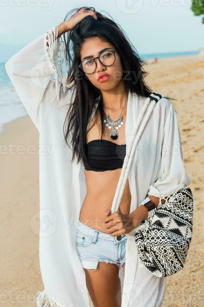 seducente asiatico donna nel nero superiore e jeans pantaloncini , spiaggia copertina su con boho Borsa in posa su spiaggia. foto